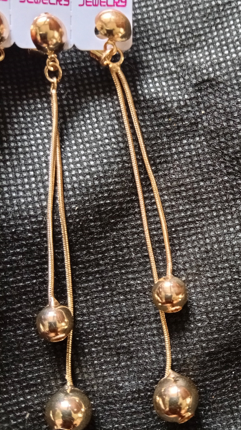 A pair of gold-toned dangle earrings with three small polished beads on each long wire, displayed on a textured black fabric background. A white retail tag is partially visible at the top right.