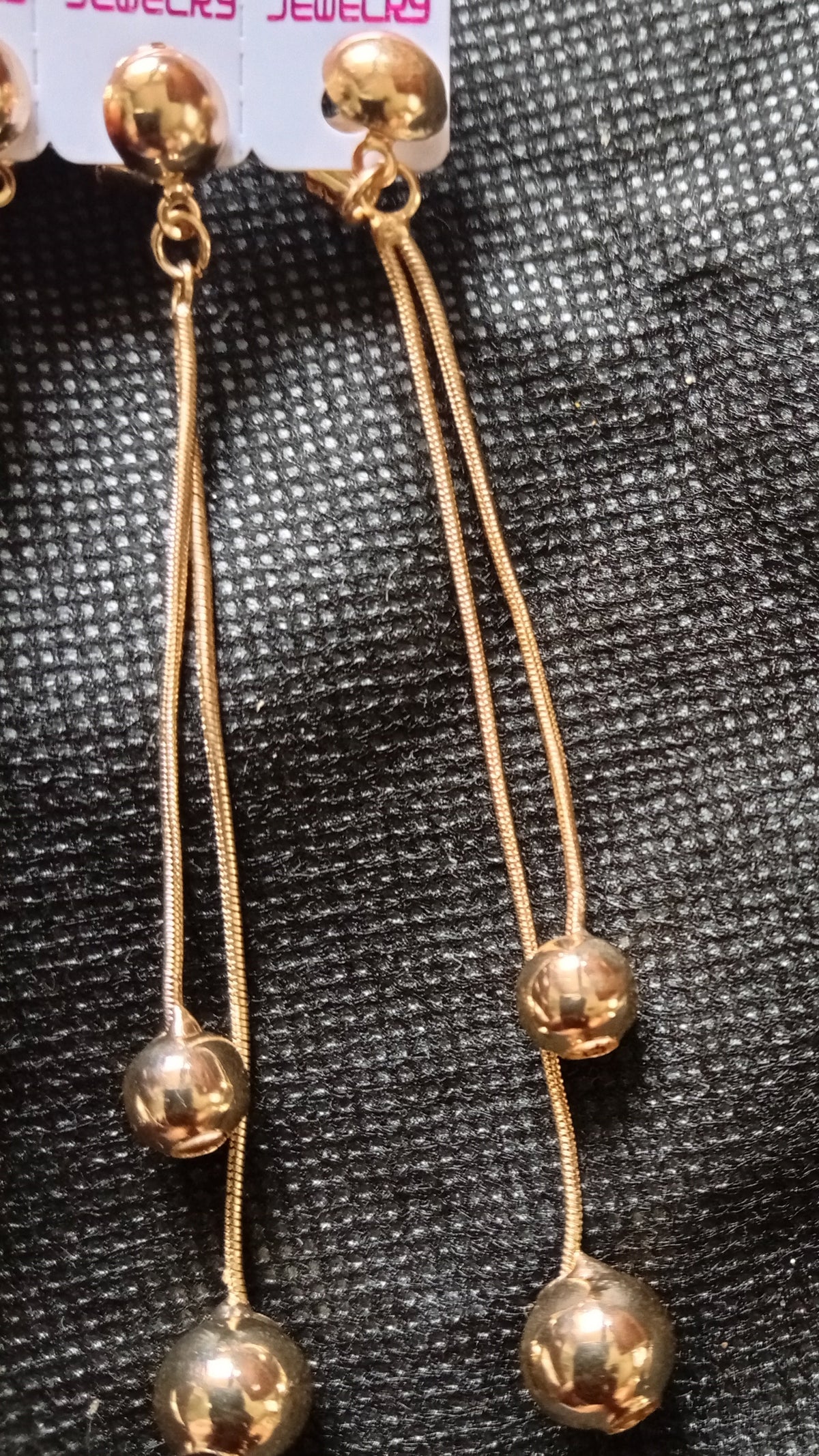 A pair of gold-toned dangle earrings with three small polished beads on each long wire, displayed on a textured black fabric background. A white retail tag is partially visible at the top right.