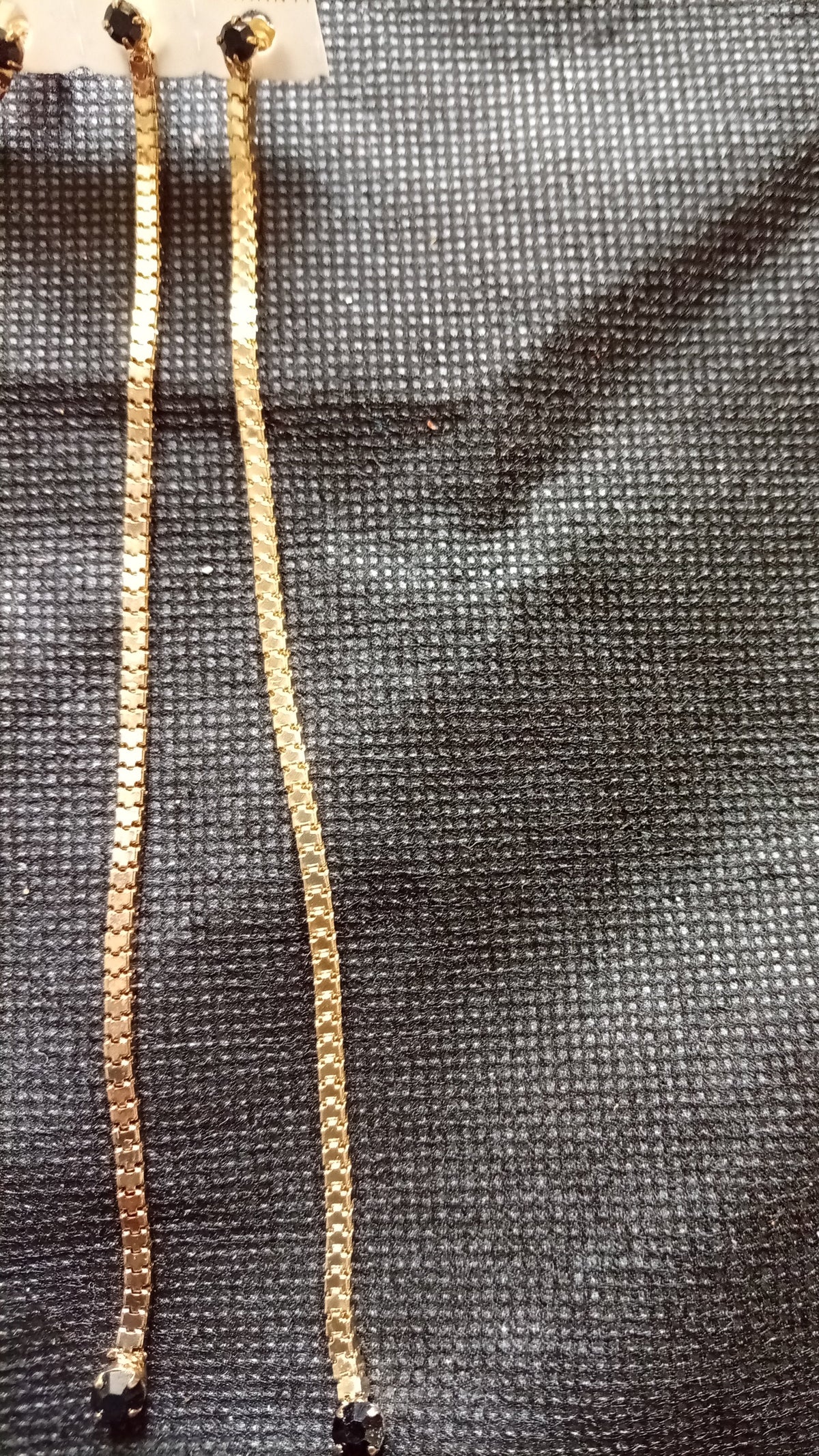 Two gold-toned earrings link cords, likely drawstring cords, laid flat on a textured dark gray fabric background.