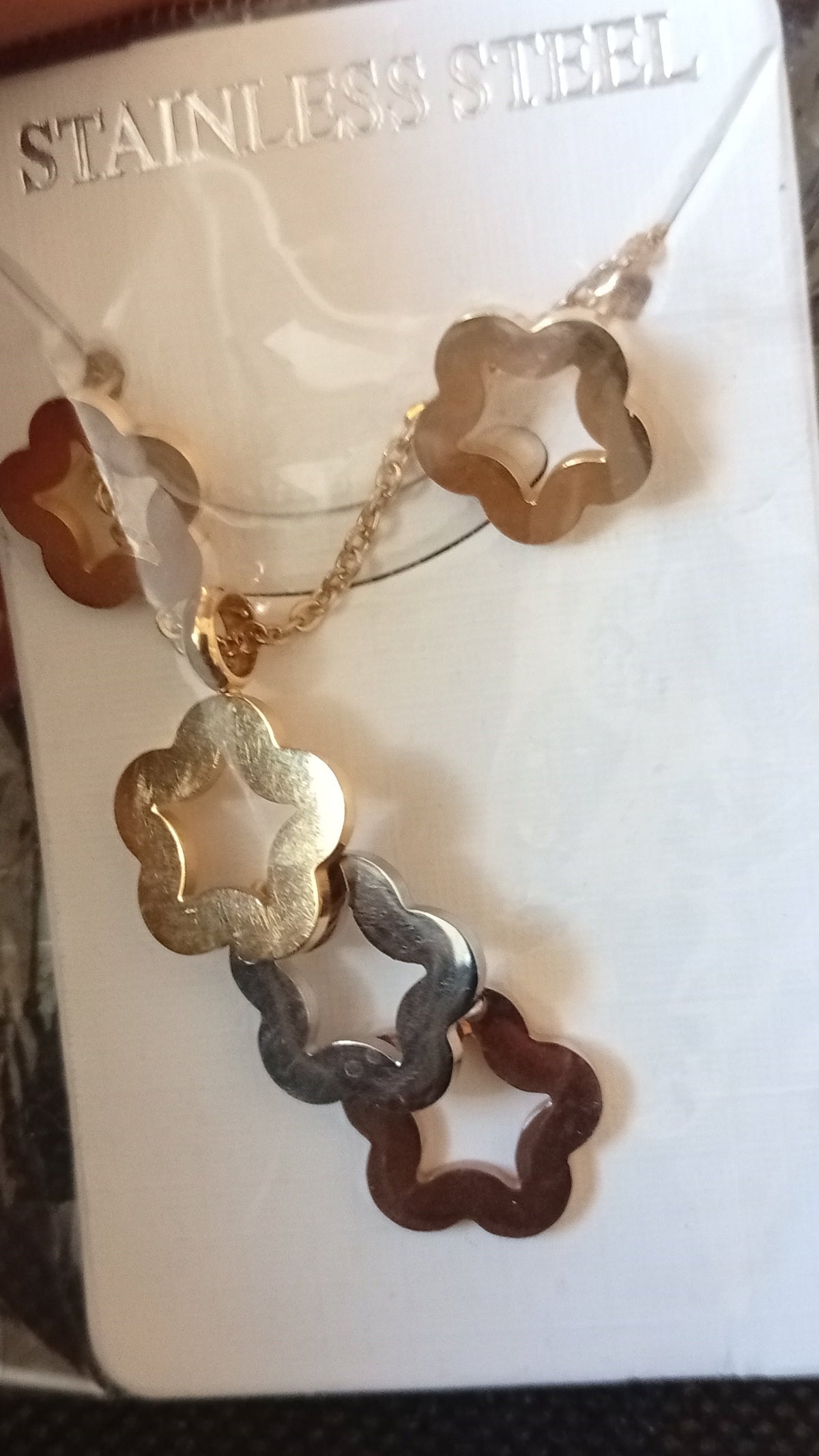 A pair of flower-shaped dangling earrings in gold and silver-tone metal, displayed on a white card inside a clear plastic bag held by a hand.
