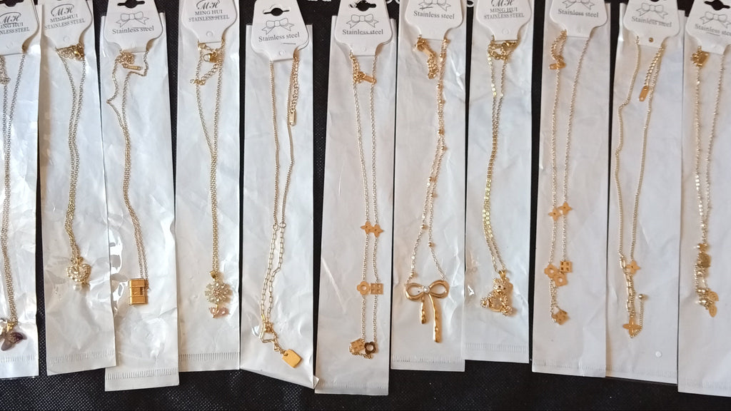 A row of ten gold necklaces with various charms, each individually packaged in white paper sleeves and displayed on a black fabric background.