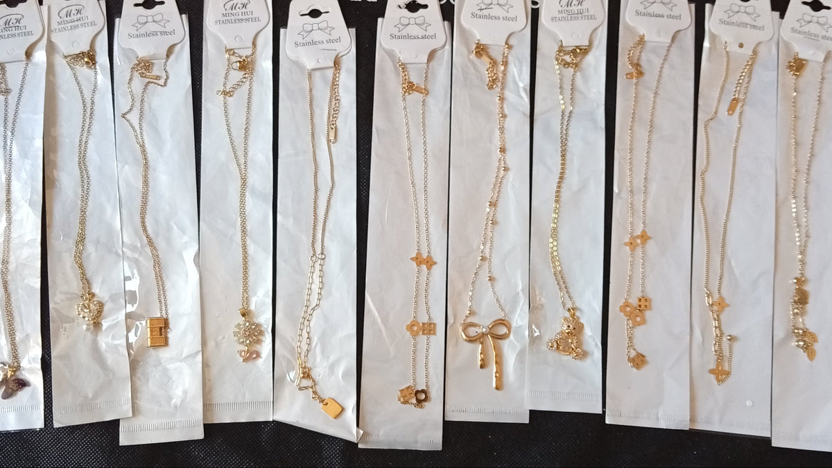 A row of ten gold necklaces with various charms, each individually packaged in white paper sleeves and displayed on a black fabric background.