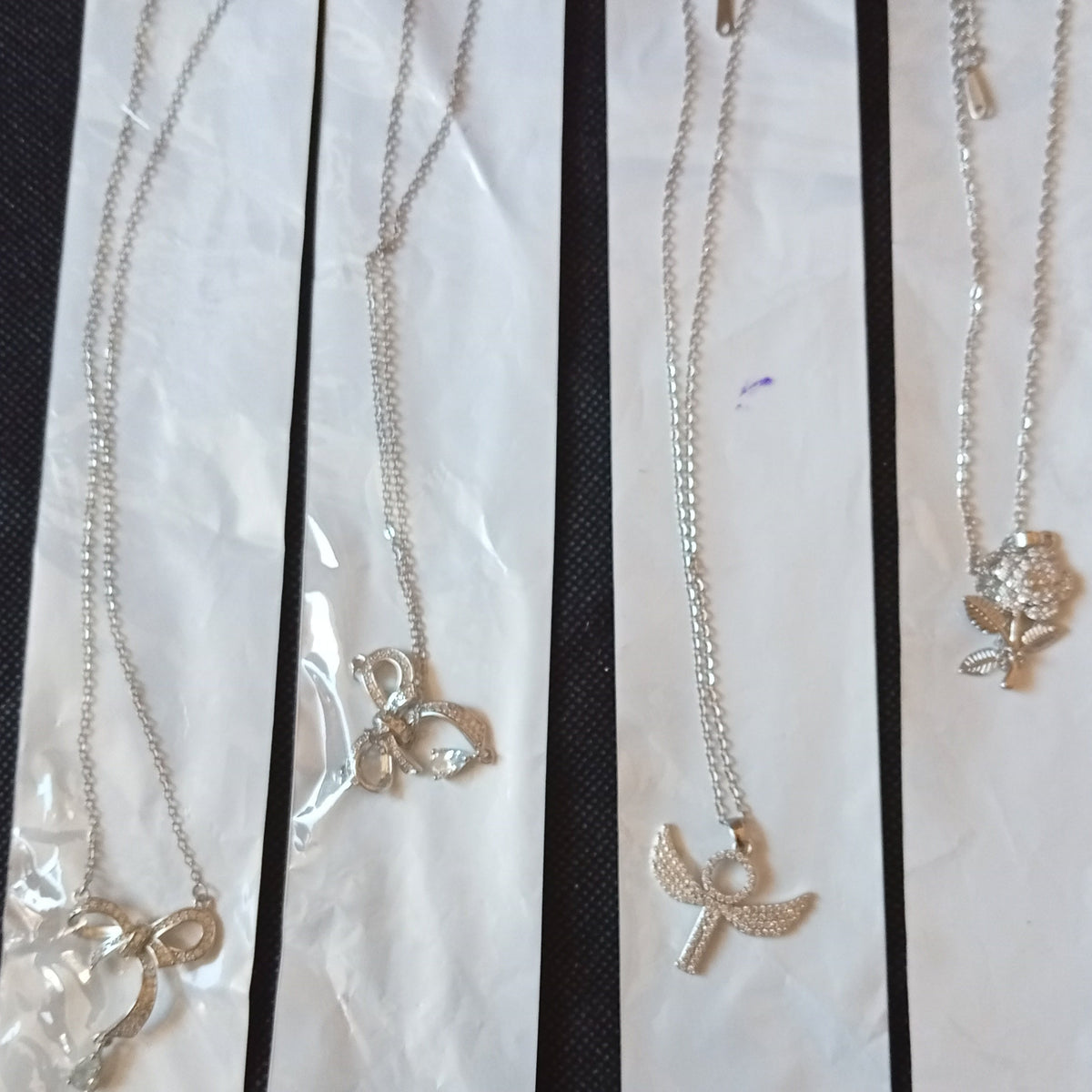 Four silver-toned chain necklaces with different small charms, each mounted on a white card labeled 'STAINLESS STEEL', laid out on a black textured fabric background.