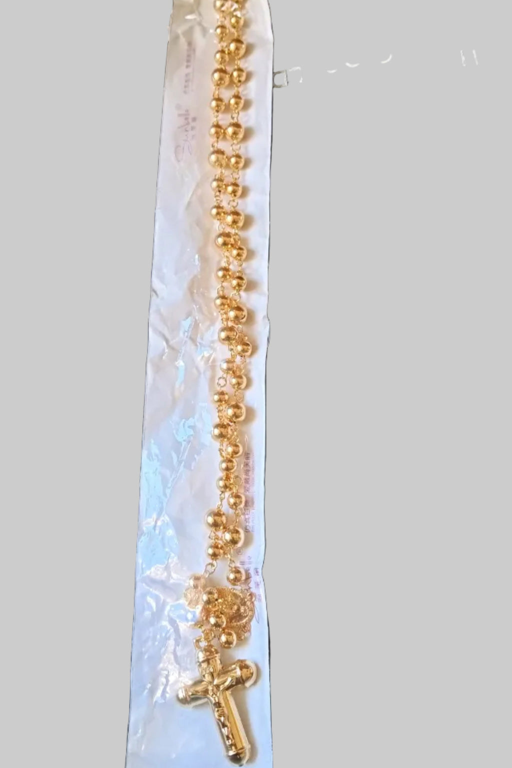 A gold-toned chain  with a cross pendant, still in its white plastic packaging, laid on a black textured surface.