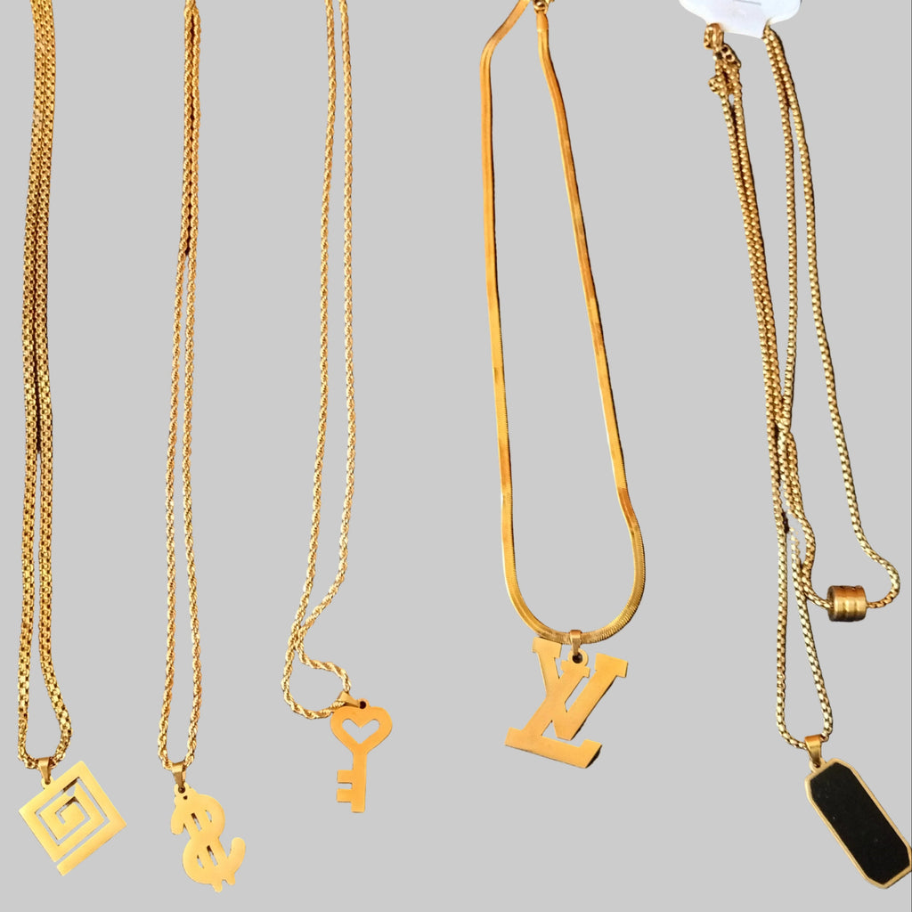 Five gold-tone chain necklaces with different pendants (a square maze, a dollar sign, a key, and a stylized 'N' or 'Z' pendant) laid out on a black textured fabric background. Two necklaces have white hang tags attached.