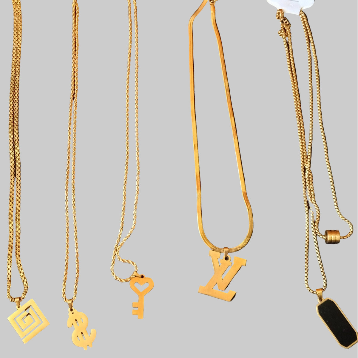 Five gold-tone chain necklaces with different pendants (a square maze, a dollar sign, a key, and a stylized 'N' or 'Z' pendant) laid out on a black textured fabric background. Two necklaces have white hang tags attached.