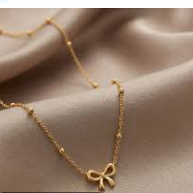 A gold bow pendant necklace with a delicate chain, displayed on draped satin fabric. The image appears to be a screenshot from a shopping gallery showing product photos, with the title 'Gold Bow Pendant Necklace' and a 'Visit' button.
