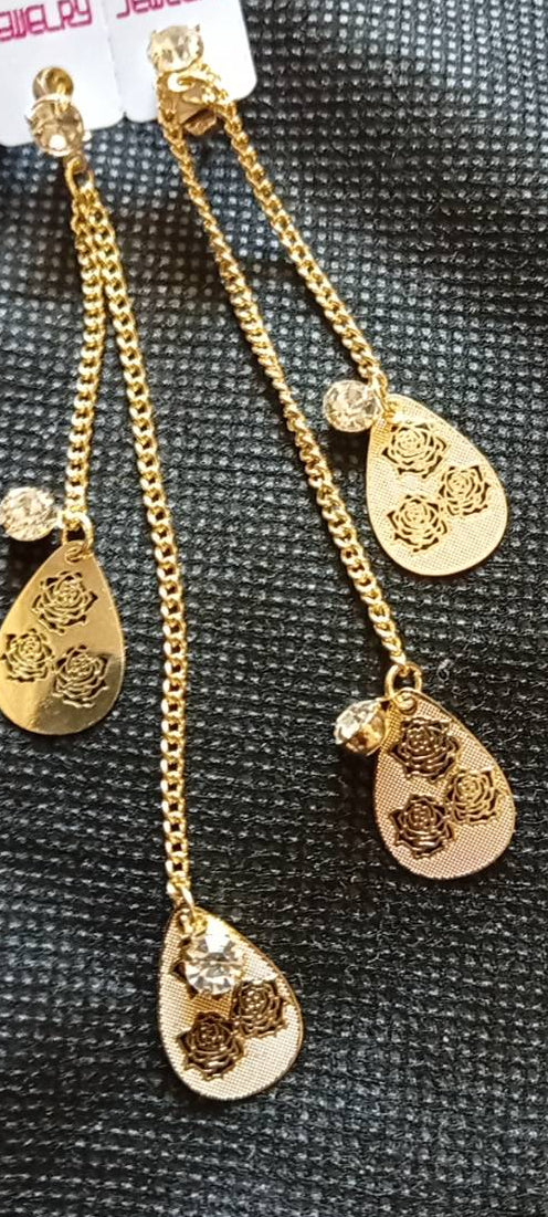 A pair of gold-toned dangle earrings with teardrop-shaped engraved charms and small clear stones, shown on a textured dark gray surface with a partial white card reading fashion jewelry.