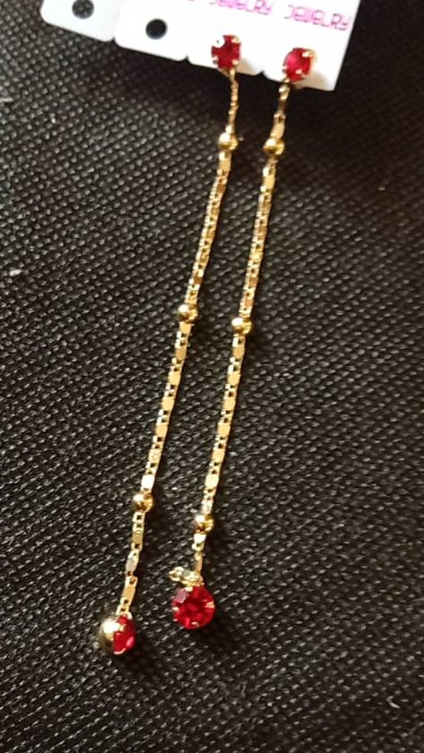 A pair of gold dangle earrings with red gemstone accents, attached to a white retail display card labeled 'Vista', placed on a dark textured surface.